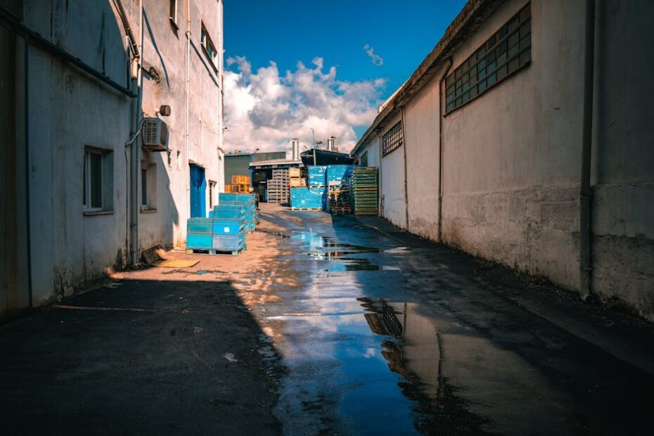 Deserted industrial alley with reflections of blue crates and sky in puddles.