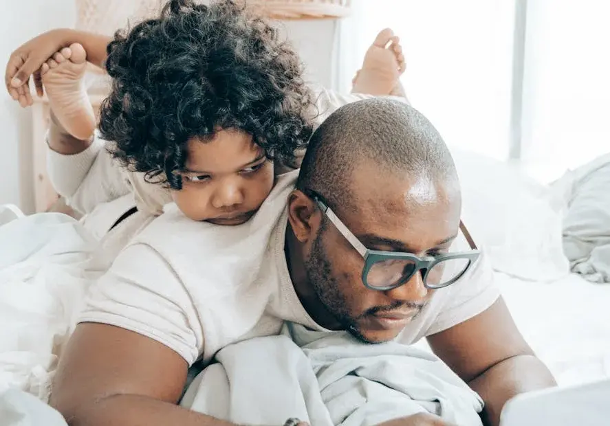 A father multitasks with his child while working on a laptop in a cozy bedroom setting.