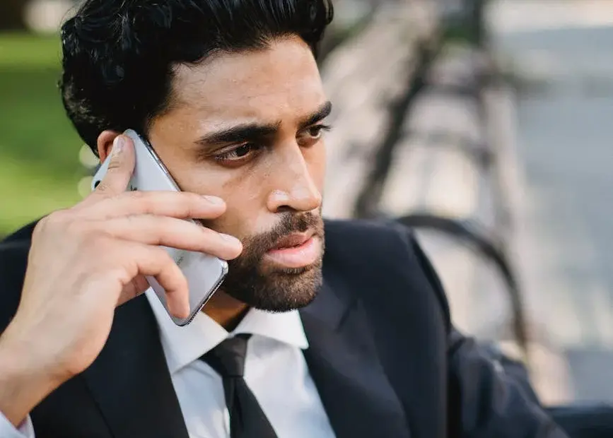 A well-dressed man making a phone call while sitting on a park bench during the day.