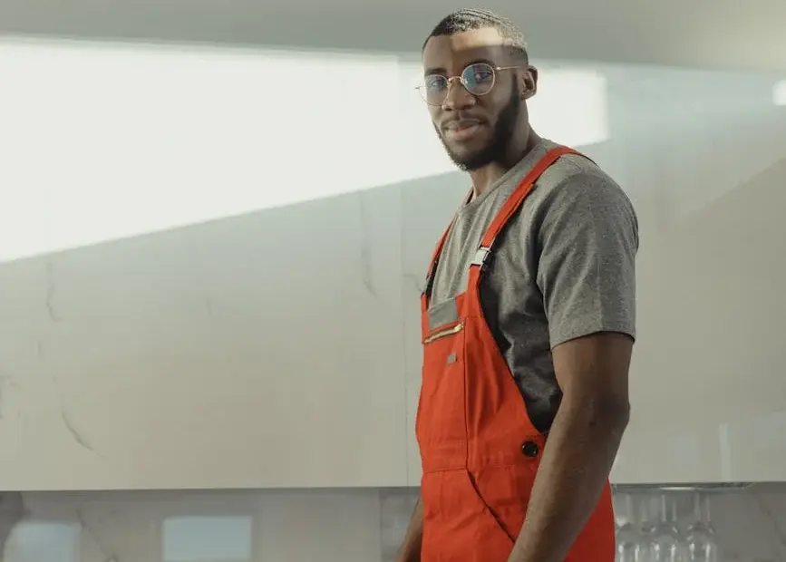 Indoor photo of a man in red overalls and glasses holding a cleaning brush, standing confidently.